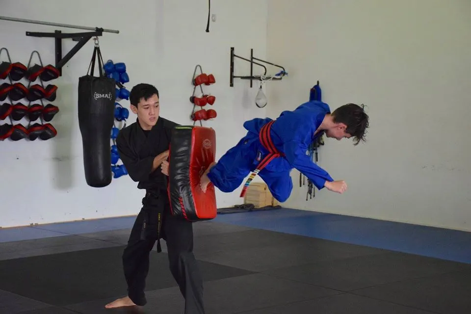 Two martial arts students practice techniques; one performs a flying kick while the other holds a focus pad.
